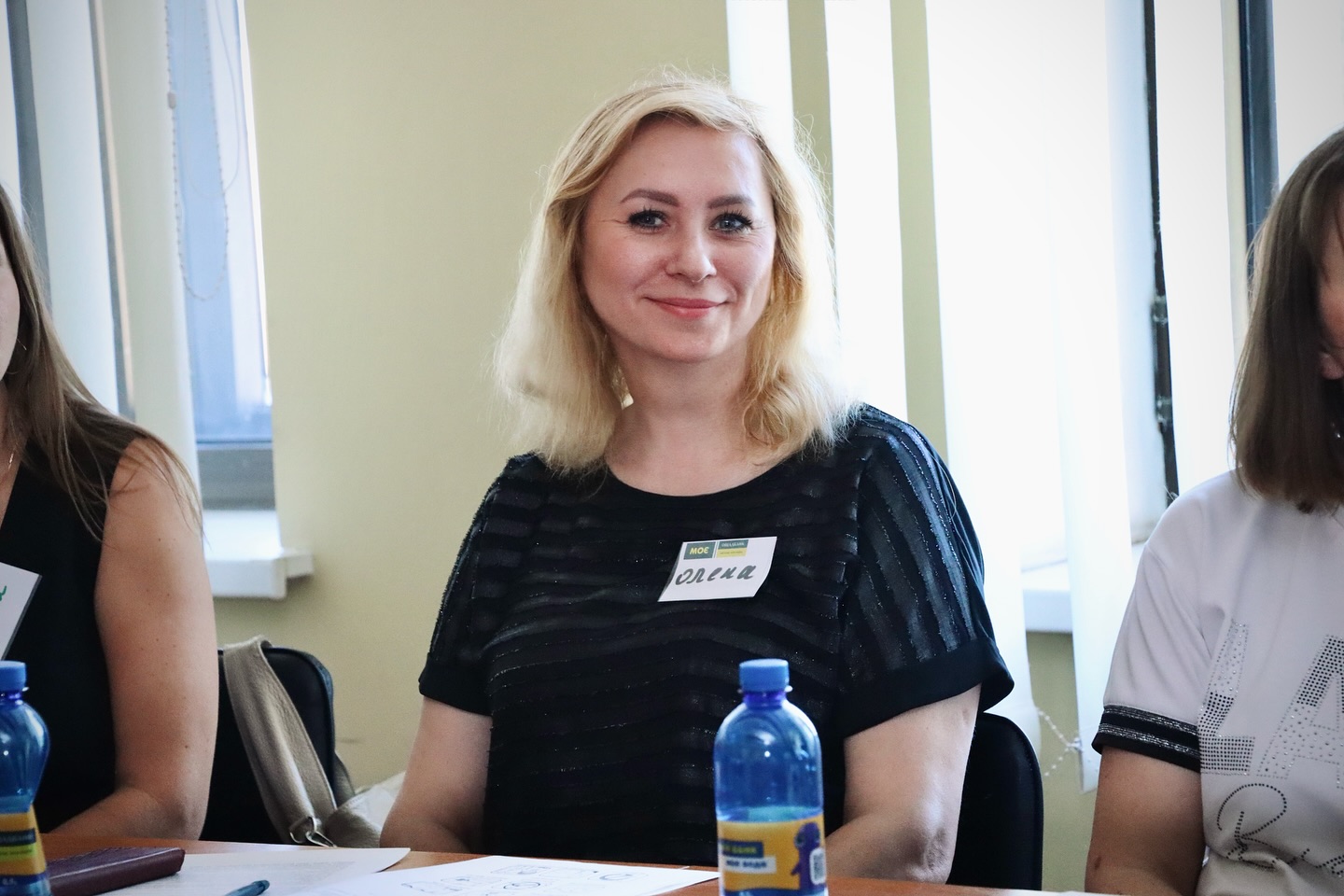 Жінка з білявим волоссям у темній блузці з бейджем 