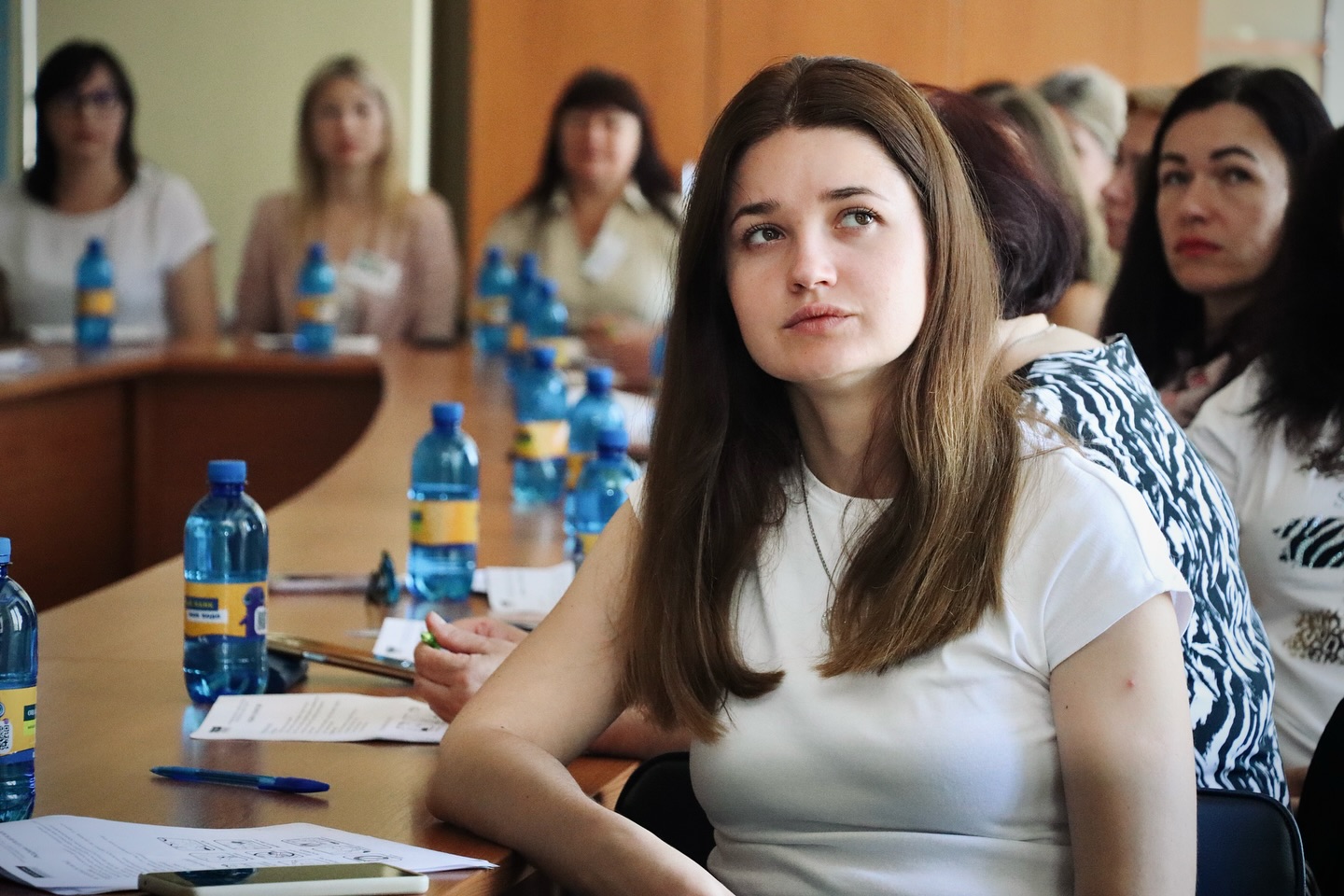 Молода жінка з довгим каштановим волоссям у білій футболці уважно слухає, сидячи за конференц-столом серед інших учасниць заходу.