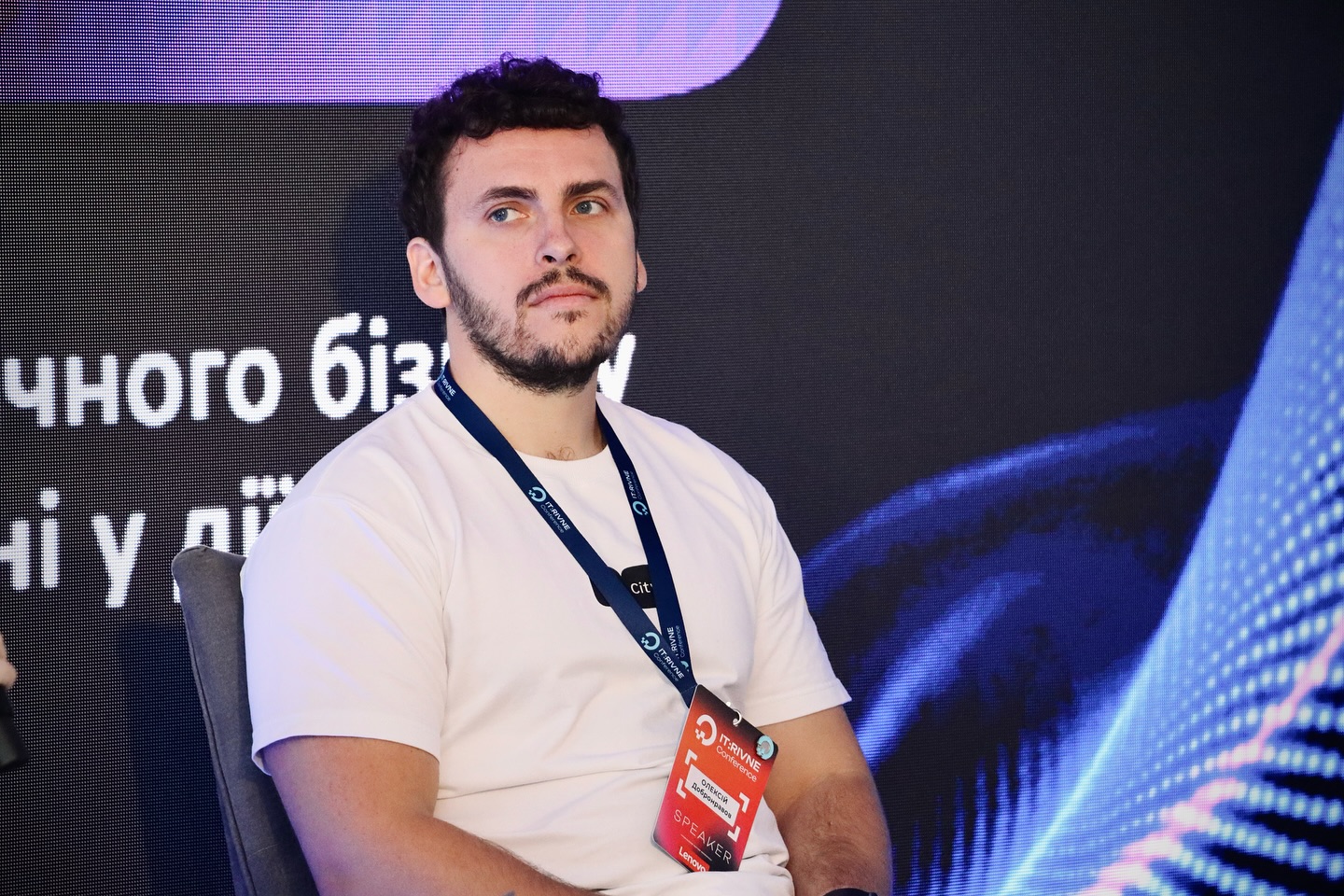 Чоловік з бородою у білому одязі сидить перед LED екраном.