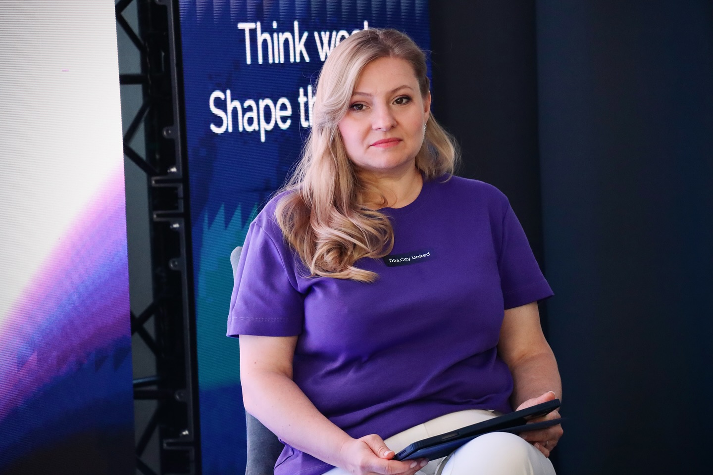 Жінка зі світлим волоссям тримає планшет та сидить перед LED екраном.