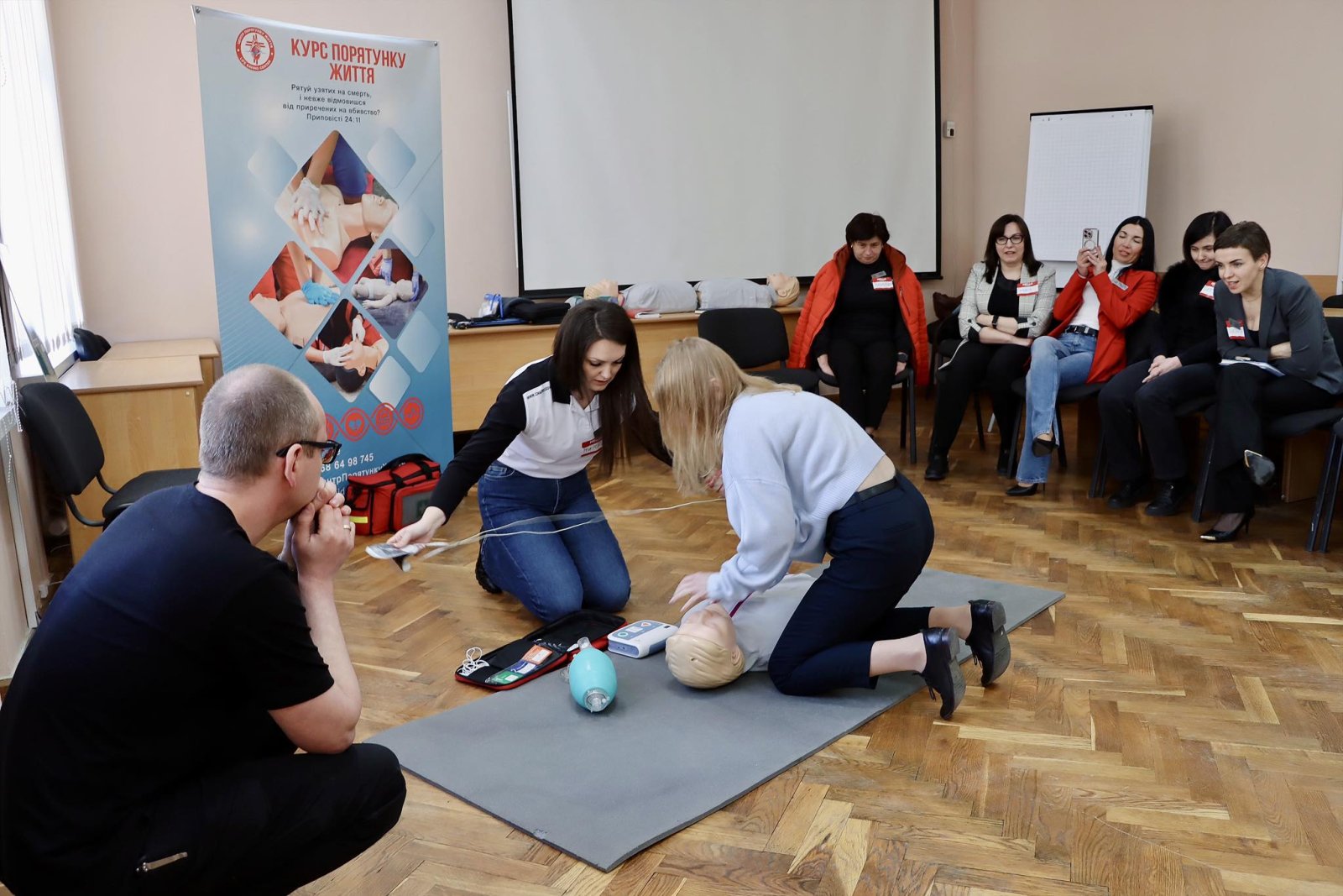 Дві жінки стоять на колінах біля манекена, збоку сидить чоловік.