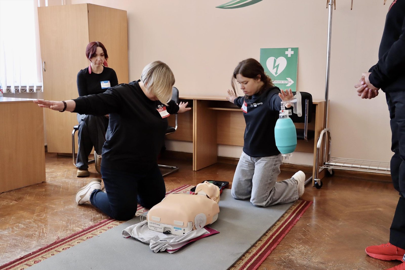 Дві жінки стоять на колінах біля манекена тримаючи руки підняті у сторони