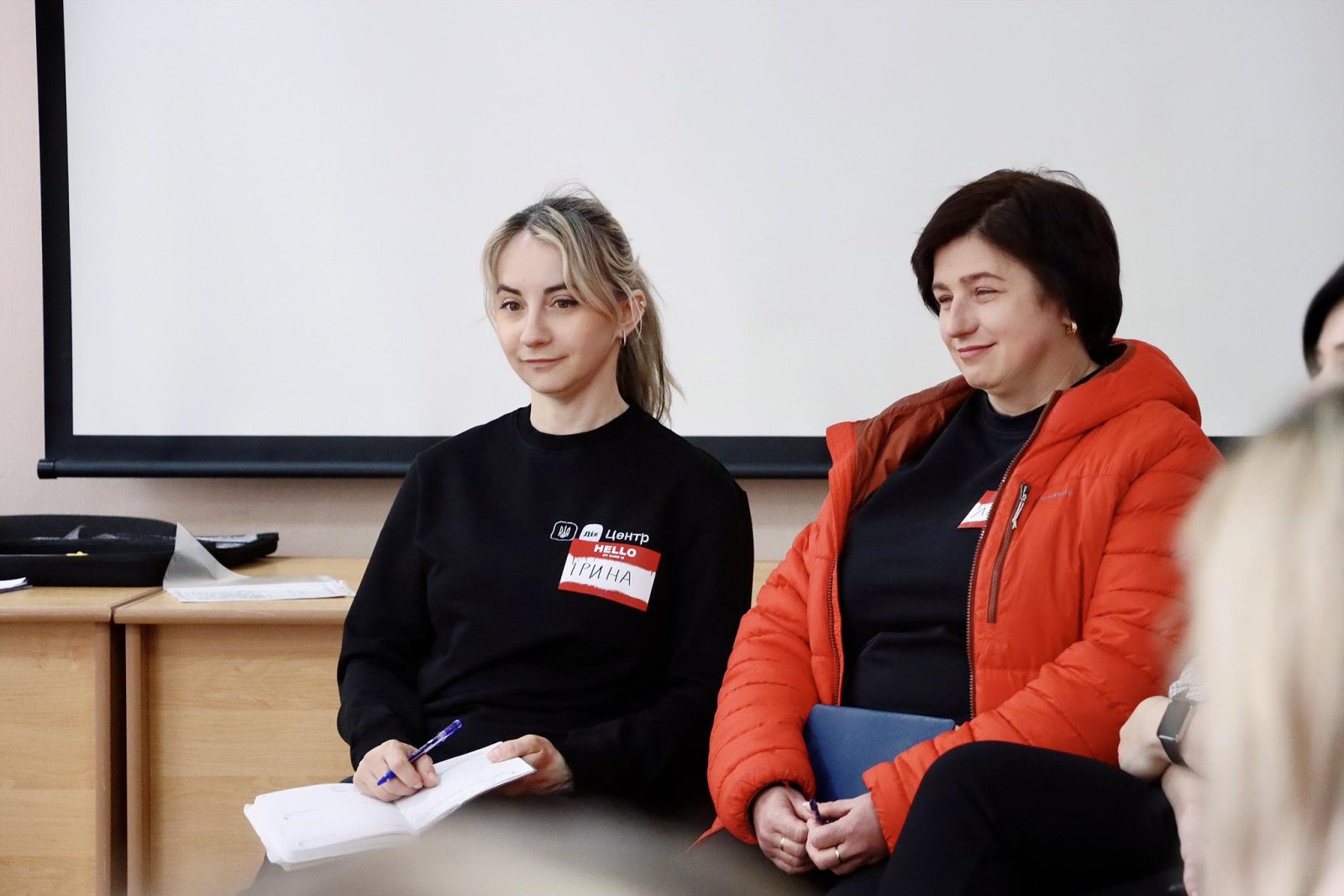 Жінка в чорній кофті та  жінка оранжевій куртці сидять у приміщенні.