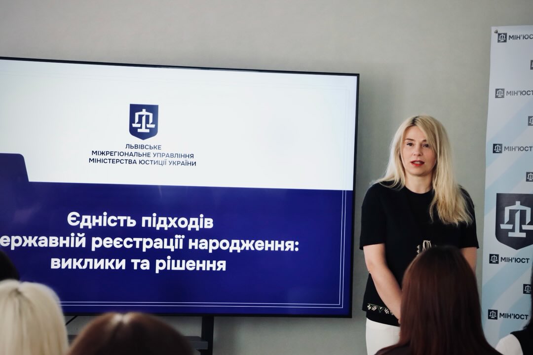 Жінка зі світлим волоссям стоїть біля екрану.