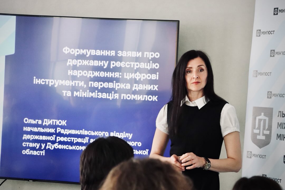 Жінка із темним волоссям стоїть біля екрану.