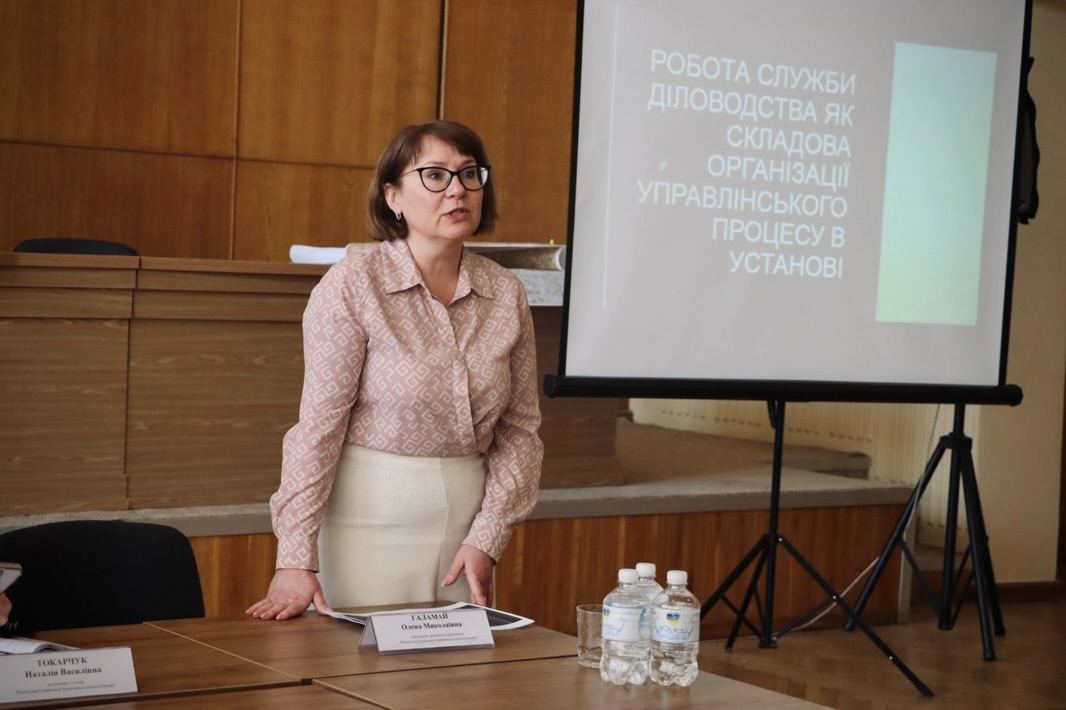 Жінка в окулярах стоїть у приміщенні.