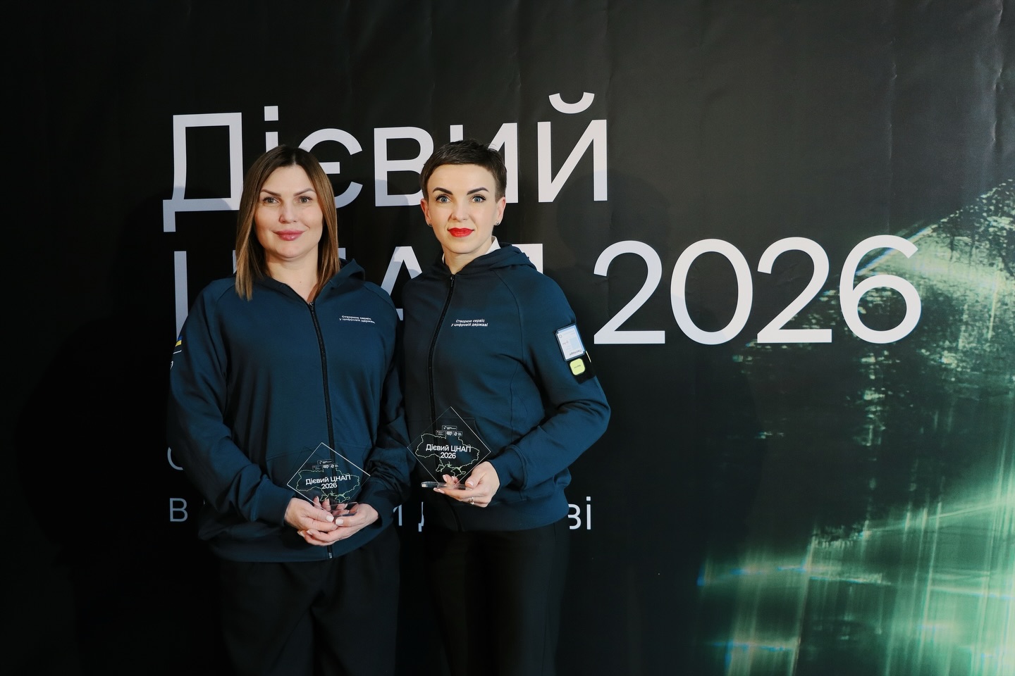 Дві жінки з нагородами позують на фоні банера.