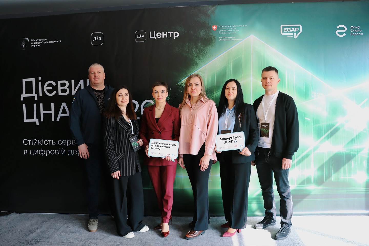 Два чоловіка та чотири жінки фоторграфуються на фоні банера.