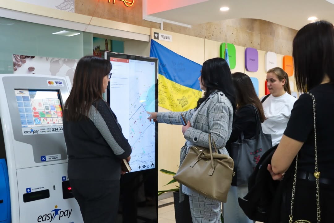 Жінка стоїть біля інформаційного терміналу, біля неї стоять інші люди.