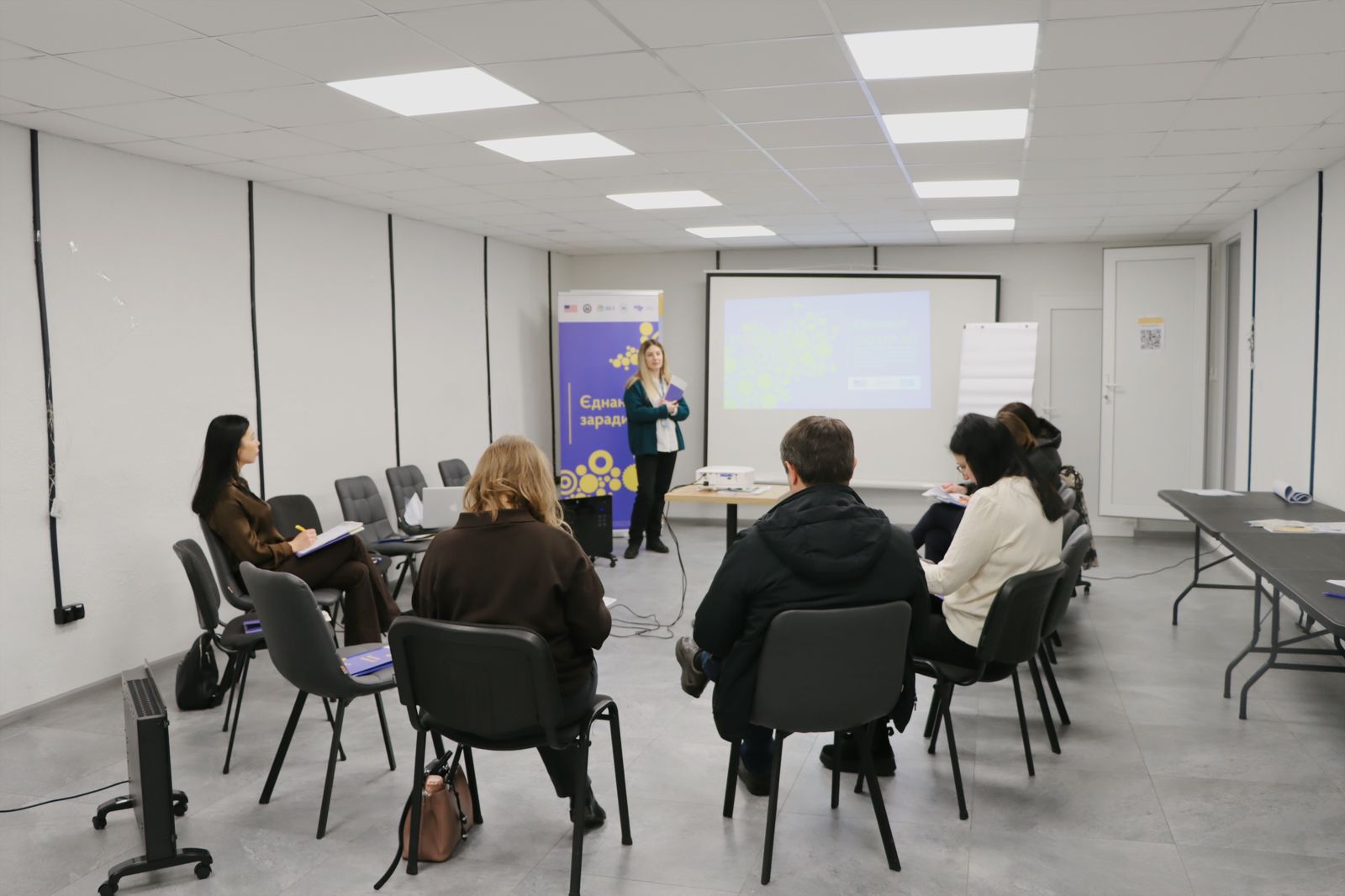 У залі проходить зустріч, жінка виступає перед групою людей, які сидять на стільцях і слухають її.