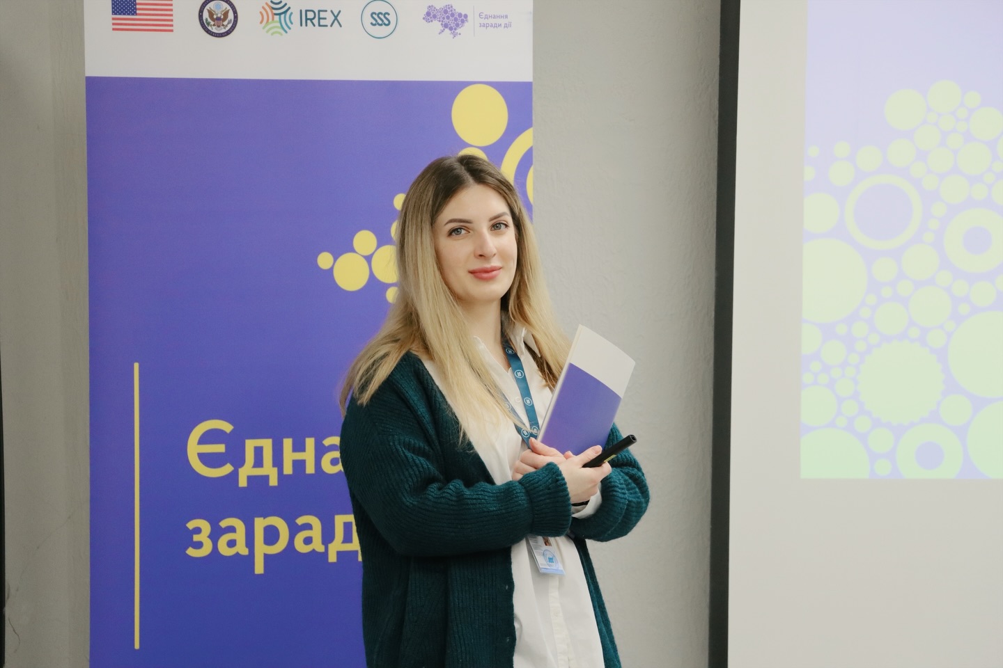 Жінка стоїть у приміщенні та посміхається.