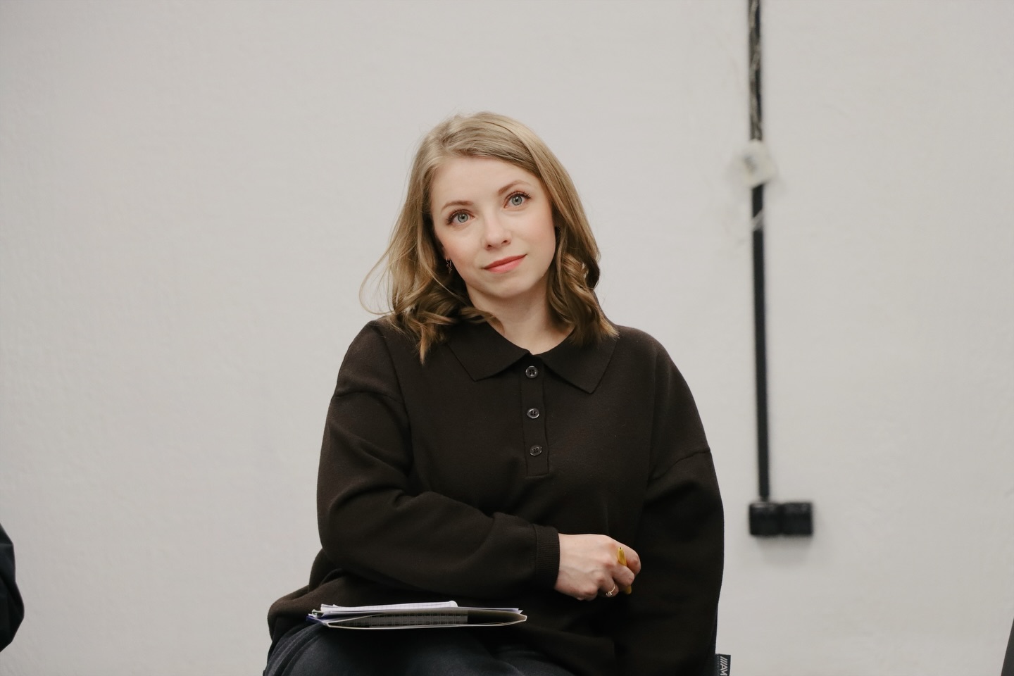 Жінка у темному одязі сидить за столом у приміщенні.