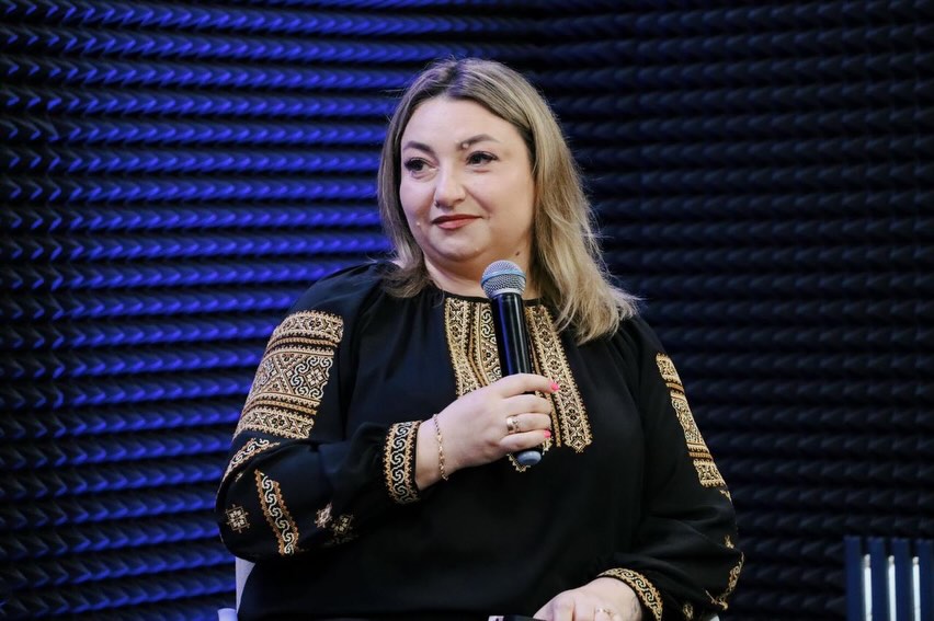 Жінка у чорній вишиванці тримає мікрофон у руках.