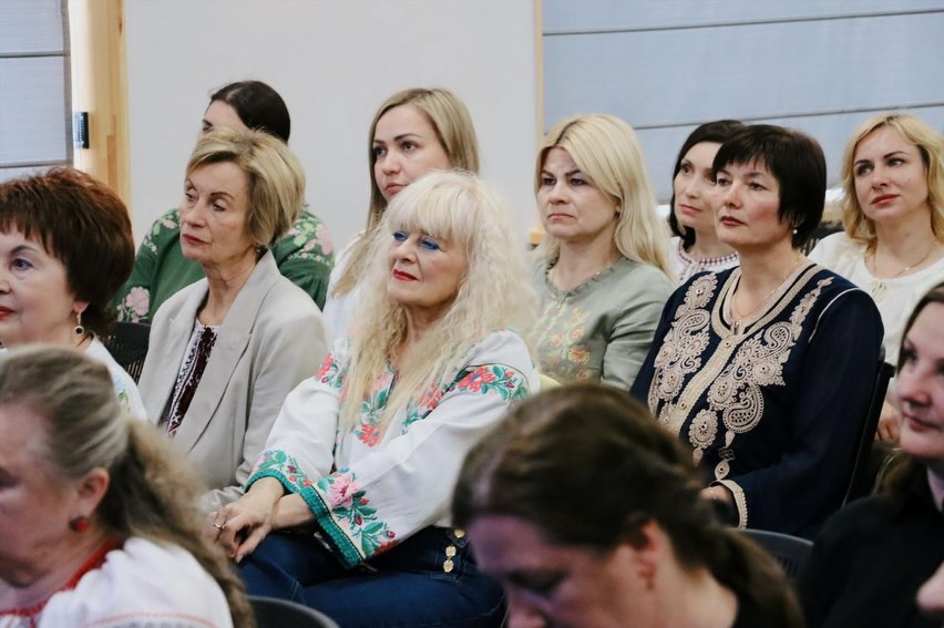 Група жінок у вишитих сорочках сидять у залі.
