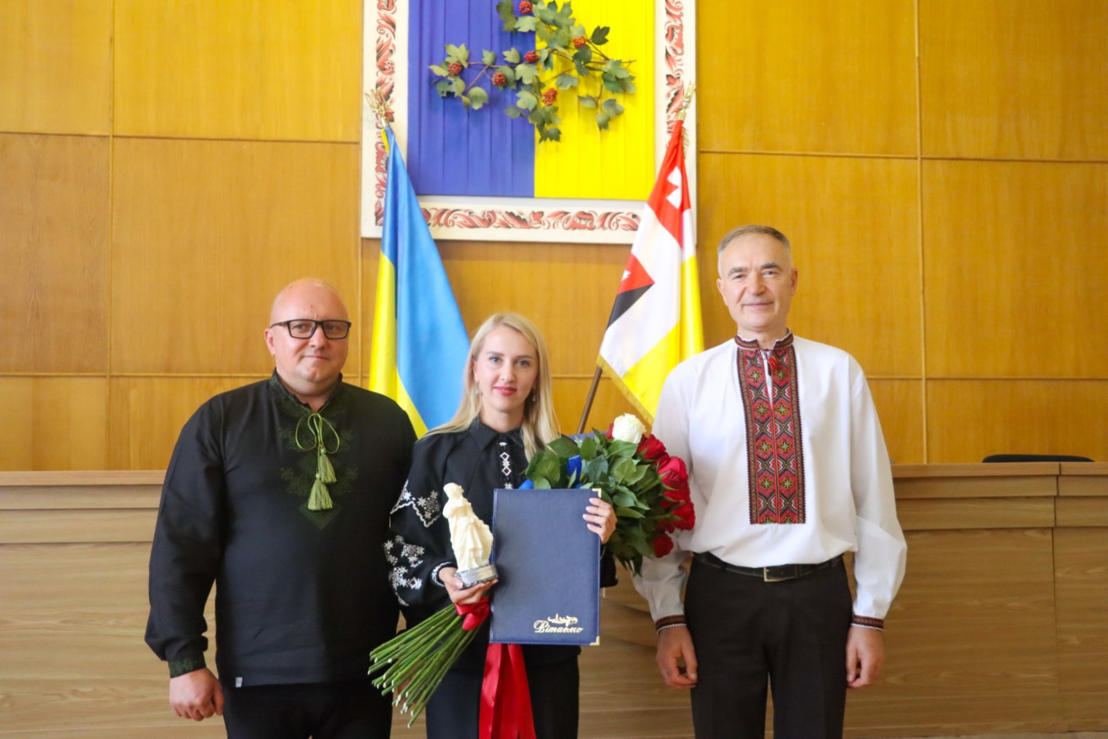 Жінка тримає квіти, біля неї стоять два чоловіка.