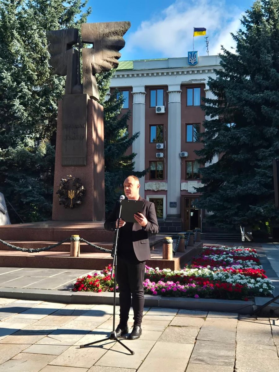 Чоговік говорить у мікрофон, позаду пам'ятник та велика будівля.
