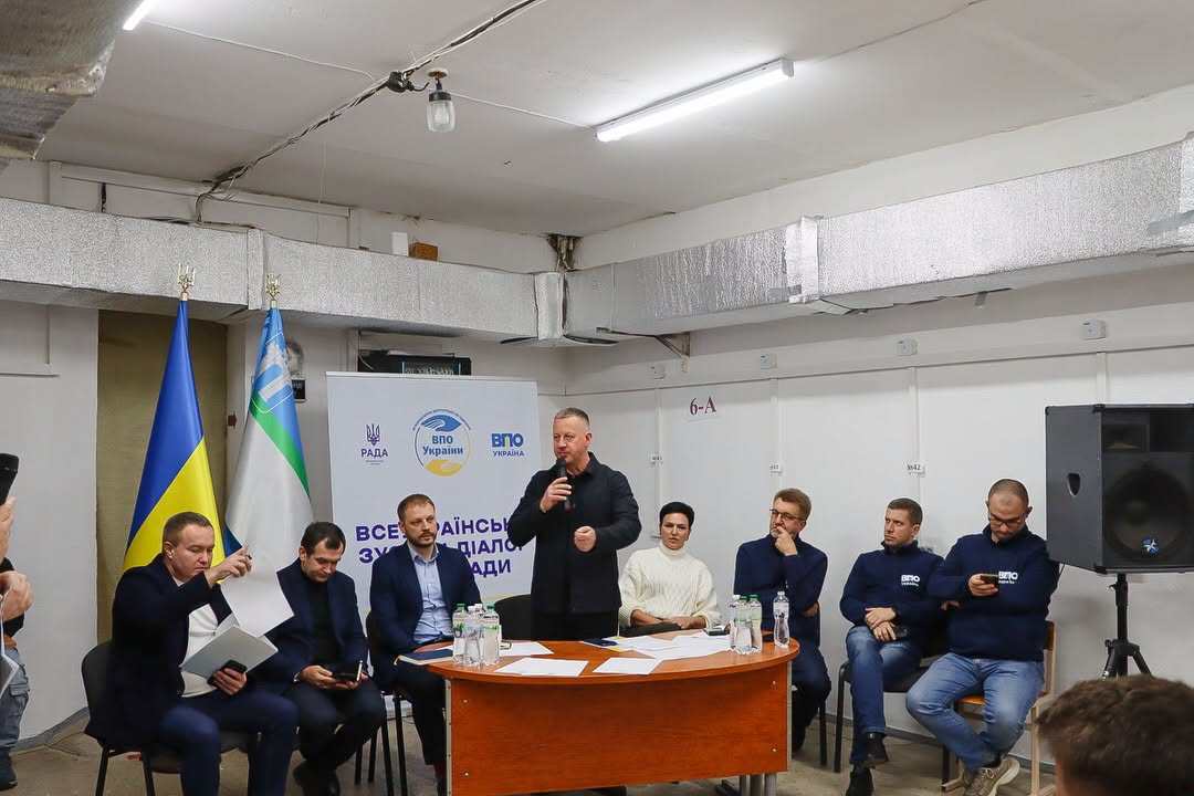 Група людей сидить у приміщенні, один чоловік стоїть і тримає мікрофон у руці.