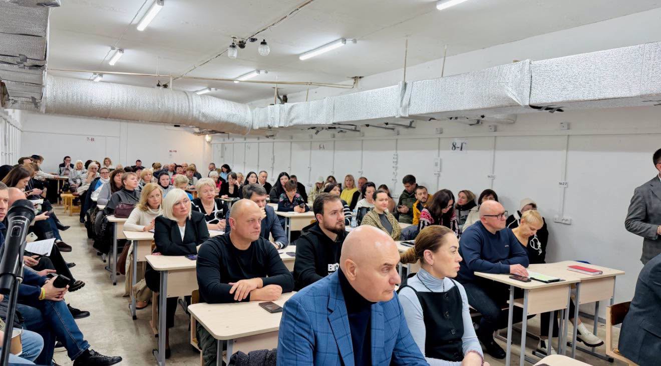 Багато людей сидить у приміщенні за партами.