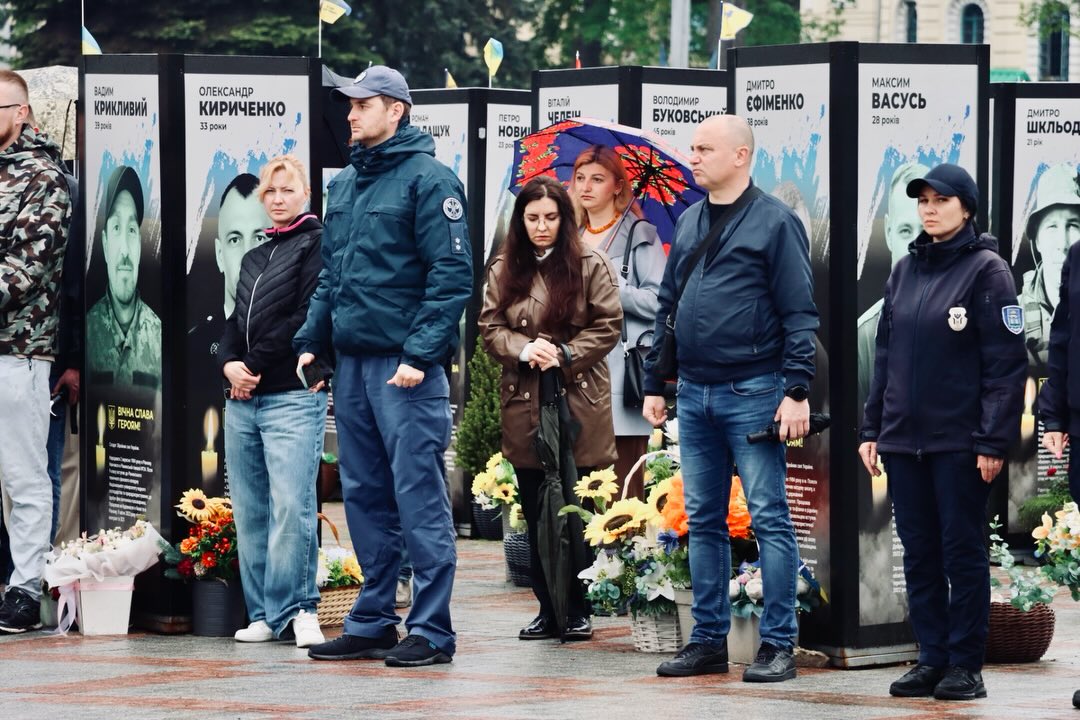 Люди стоять надворі, позаду портрети.