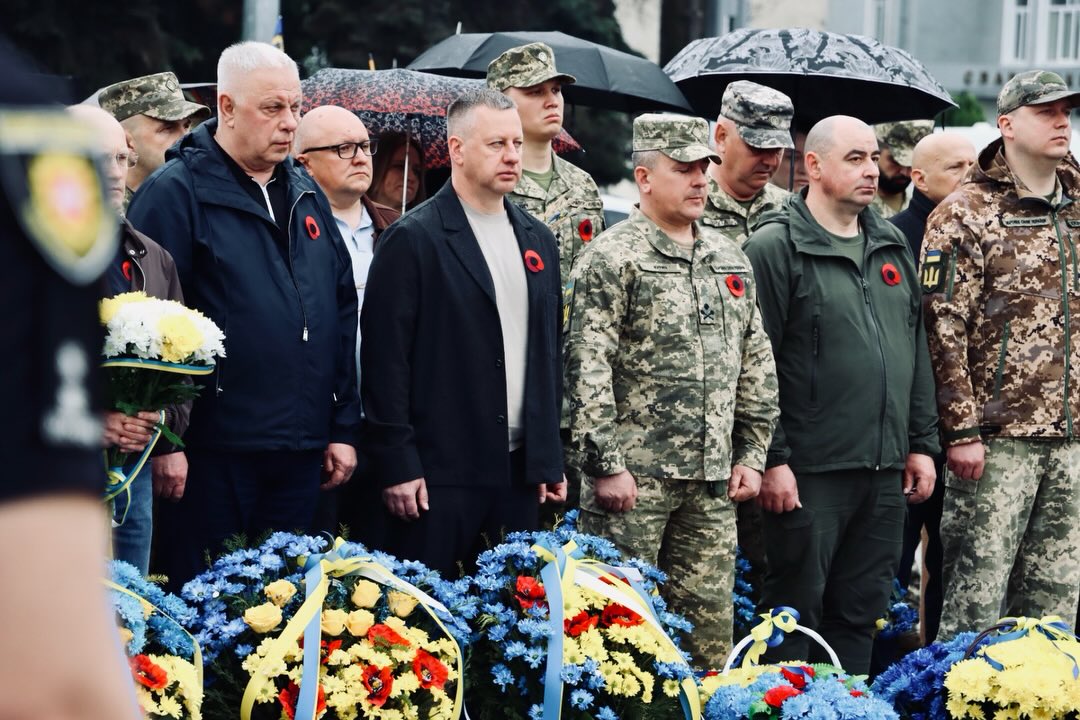 Чоловіки з червоними маками на одязі, перед ними стоять корзини з квітами.