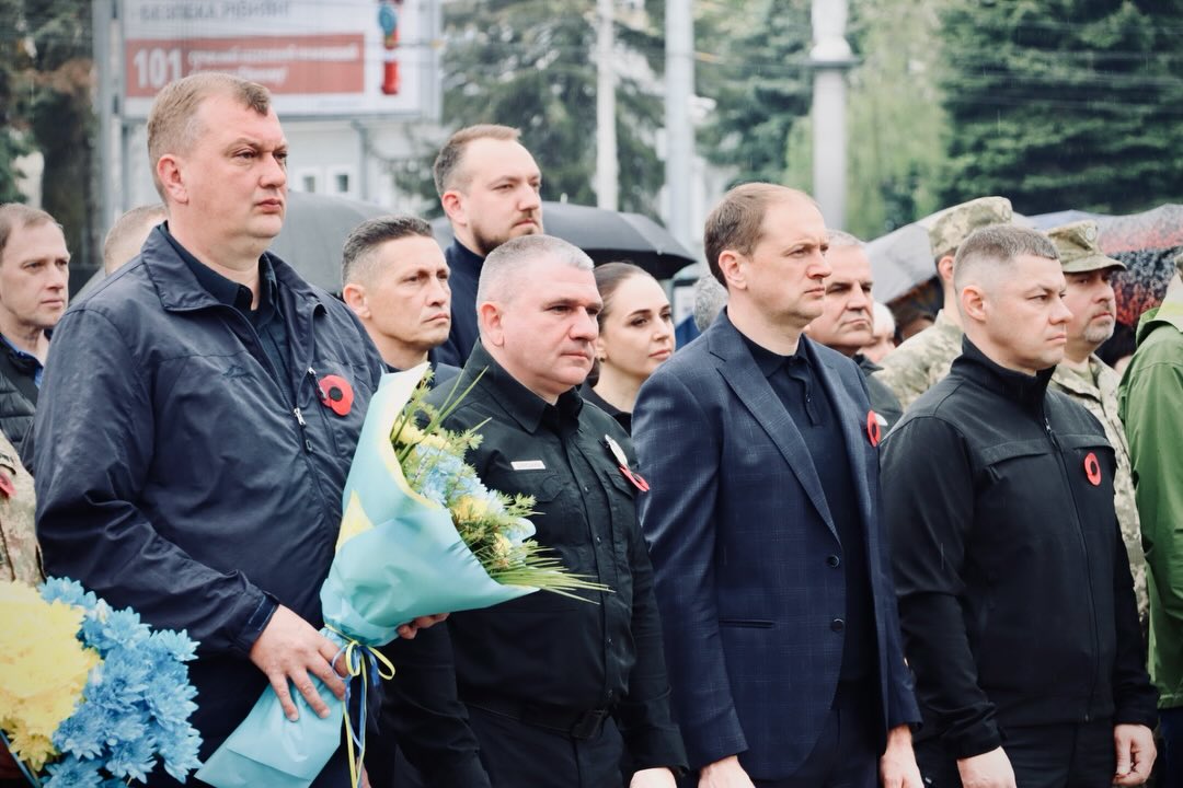 Група людей перебуває надворі, чоловік тримає букет у руках.