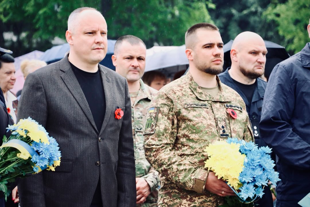 Двоє військових та декілька чоловіків у чорному одязі стоять надворі.
