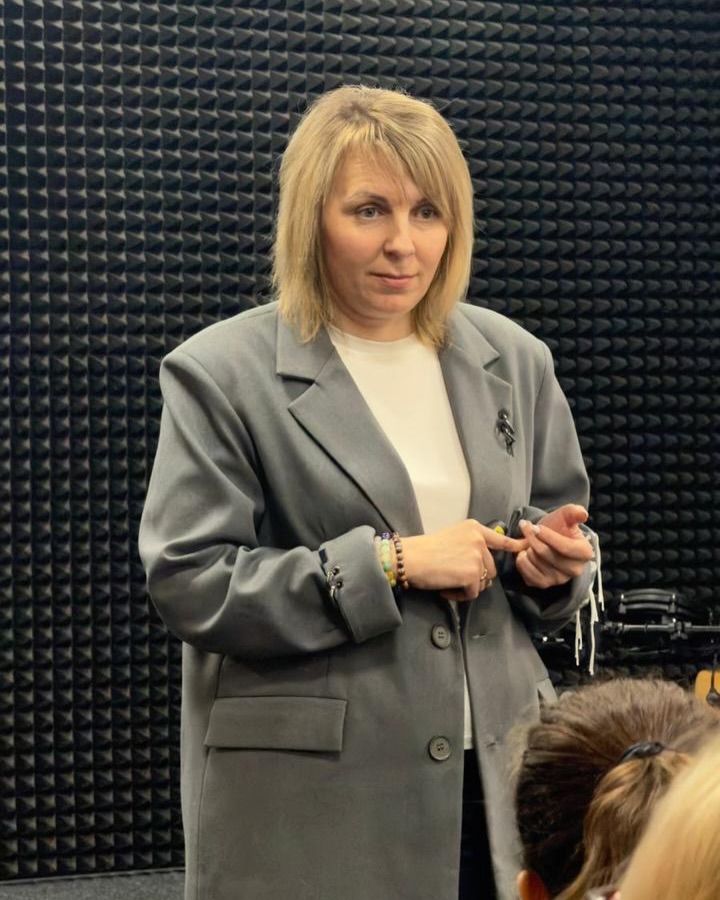 Жінка у сірому піджаку на фоні стіни.