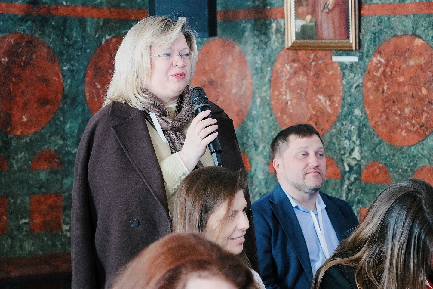 Жінка зі світлим волоссям та в окулярах тримає мікрофон у руках.