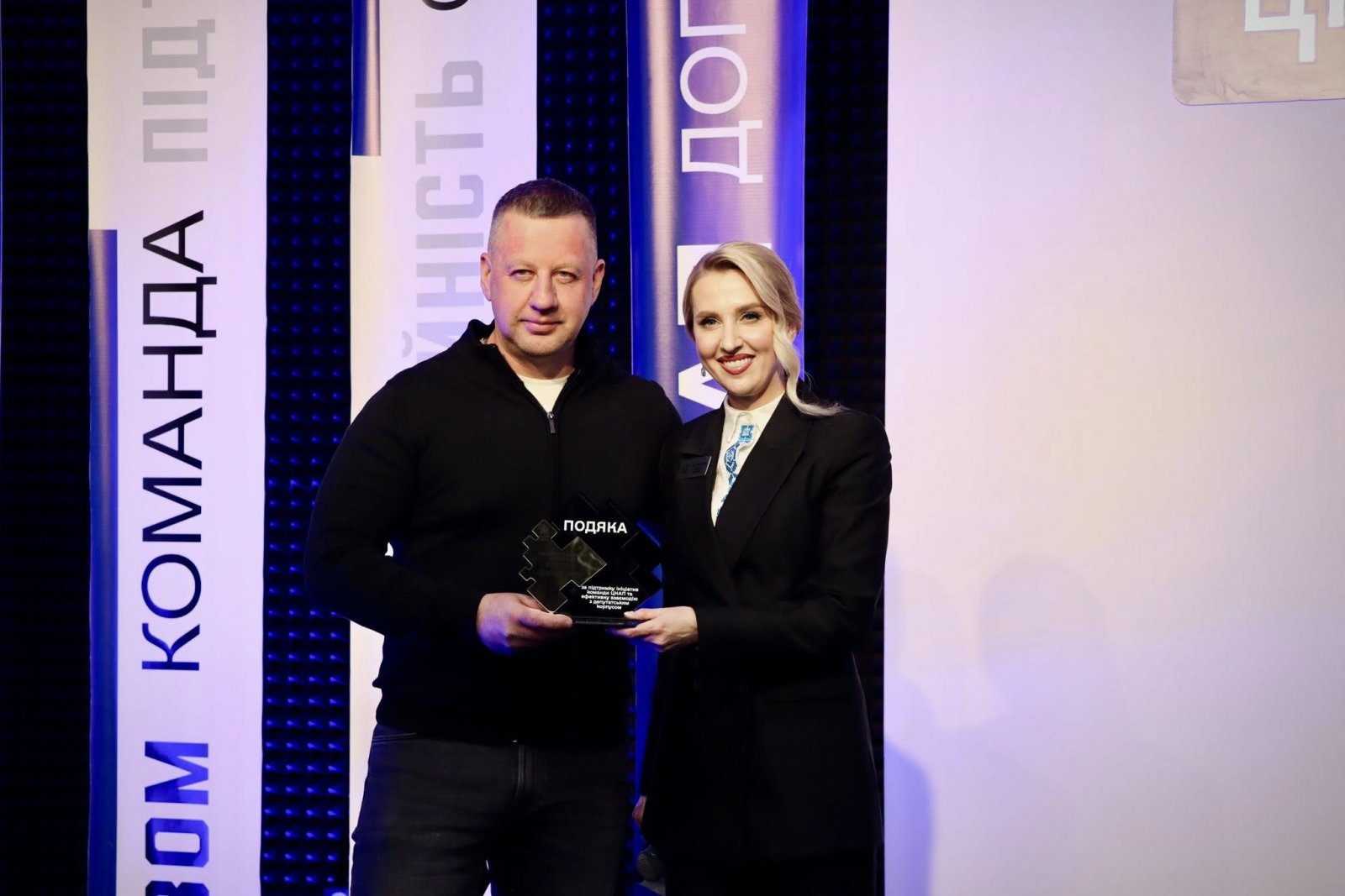 Жінка в чорному піджаку та чоловік тримають подяку