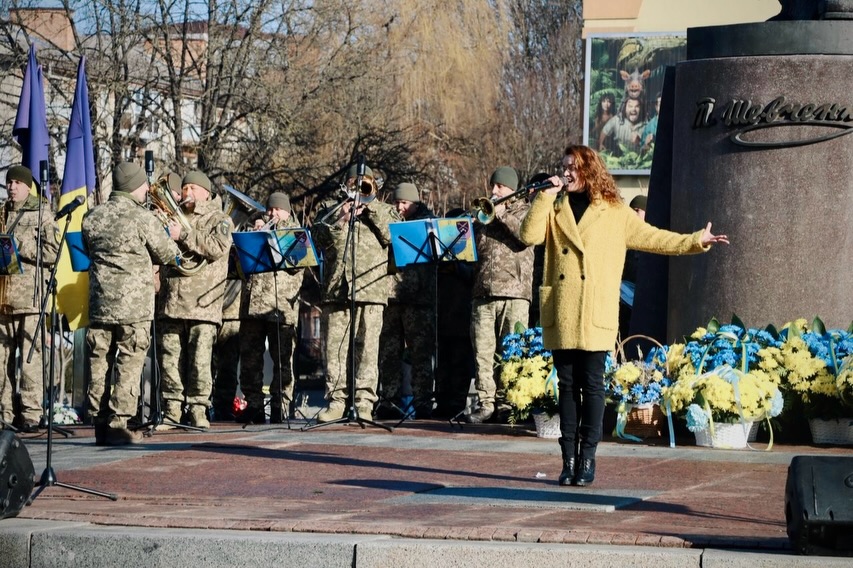 Жінка співає біля пам'ятника, збоку стоять чоловіки у військовій формі.