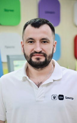 Чоловік із короткою темною стрижкою та бородою у білій футболці з логотипом.