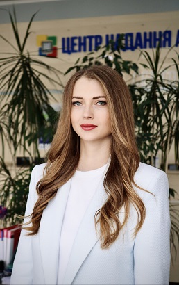 Жінка з довгим світло-русявим волоссям, у світло блакитному піджаку та білій футболці.
